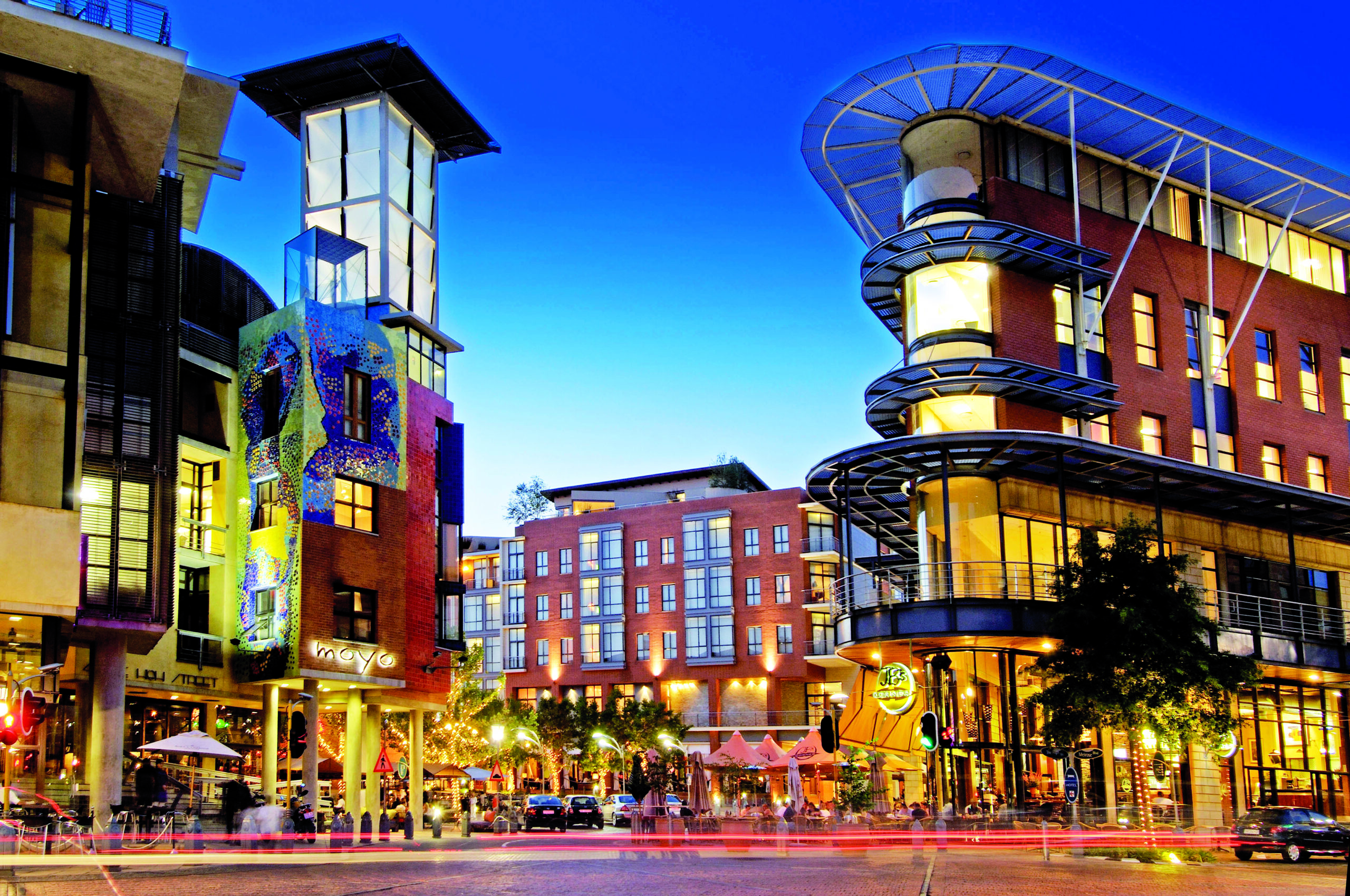 Vibrant city street at dusk with modern buildings and lively outdoor dining.